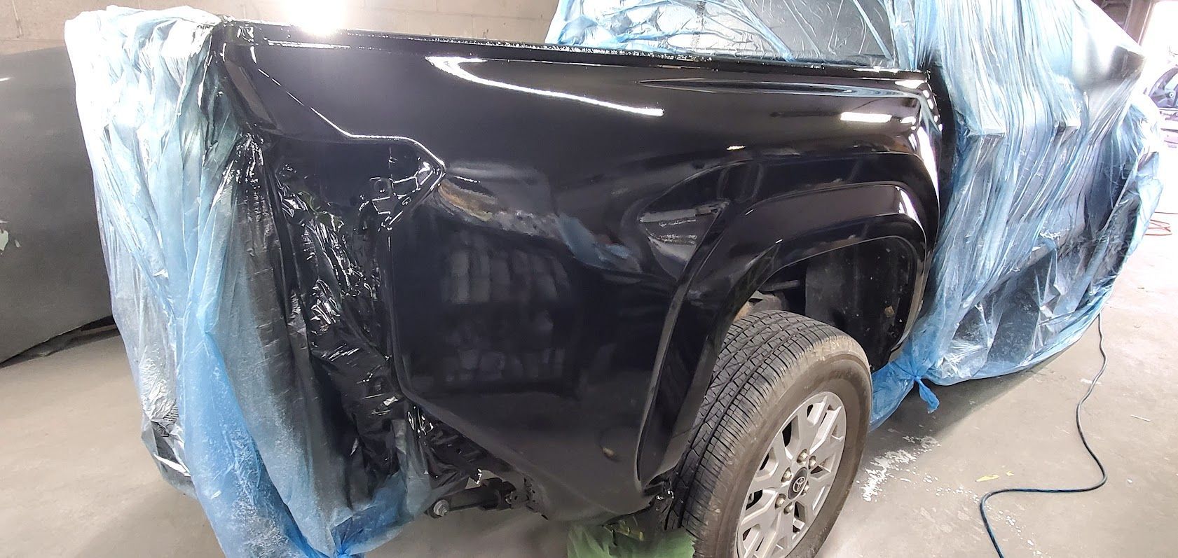 Dented black car body panel with a wheel, partially covered by blue tarp in a workshop