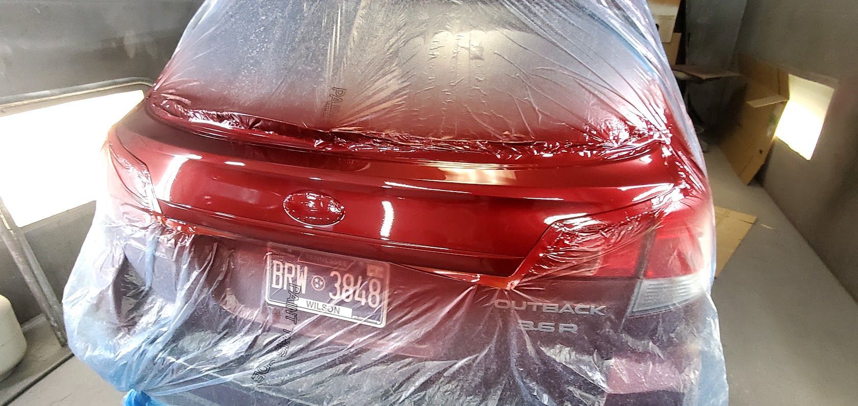 Red sedan wrapped in clear plastic inside a covered spray booth or repair bay