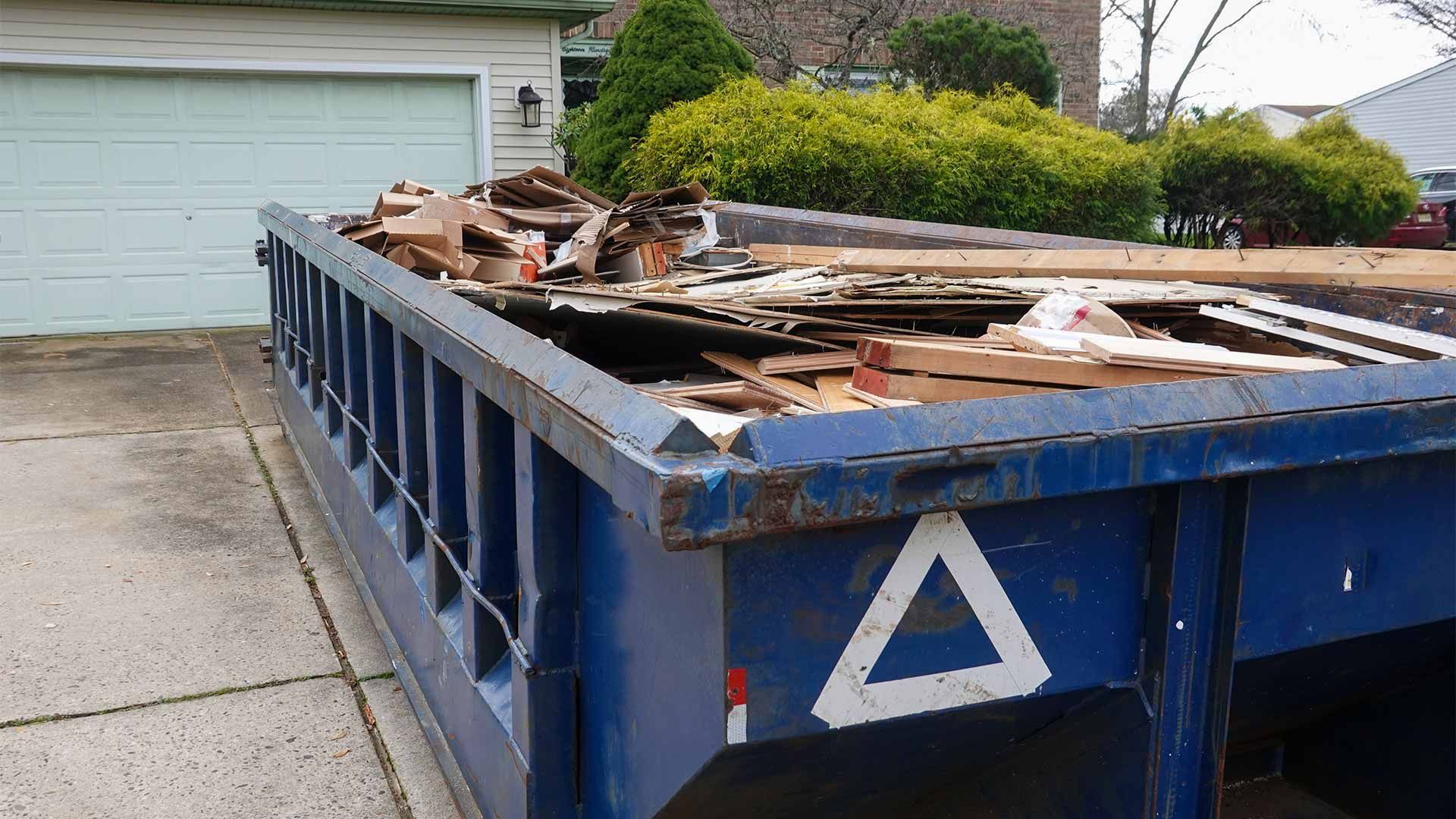 A blue dumpster filled with wood is parked in front of a house.