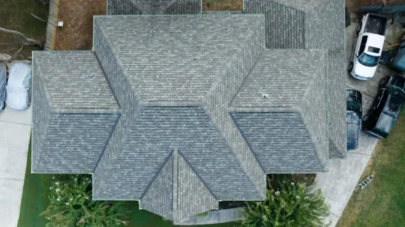 Overhead view of a house with a gray shingle roof, surrounded by cars on a driveway and trees.