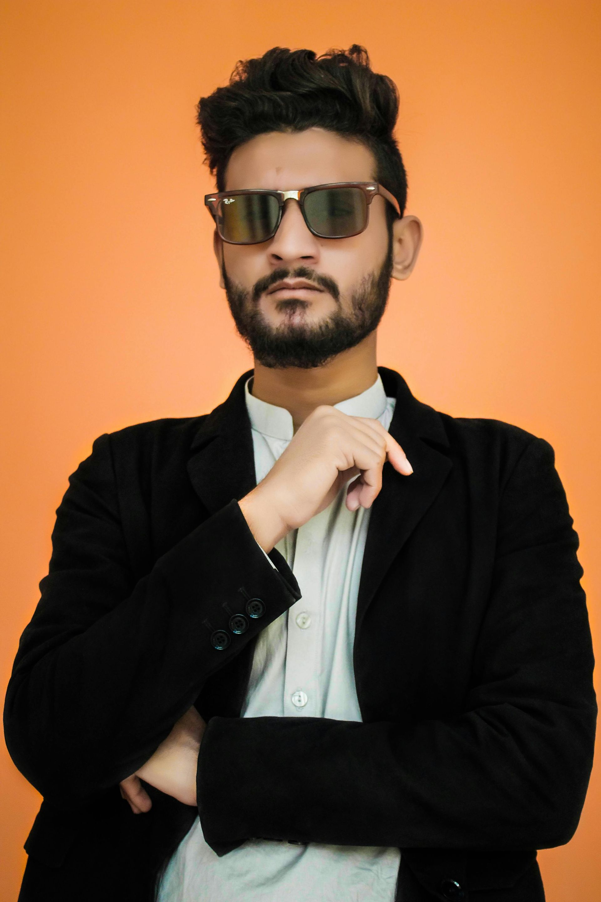 Man in sunglasses and blazer, looking at the camera, posing with crossed arms. Orange background.