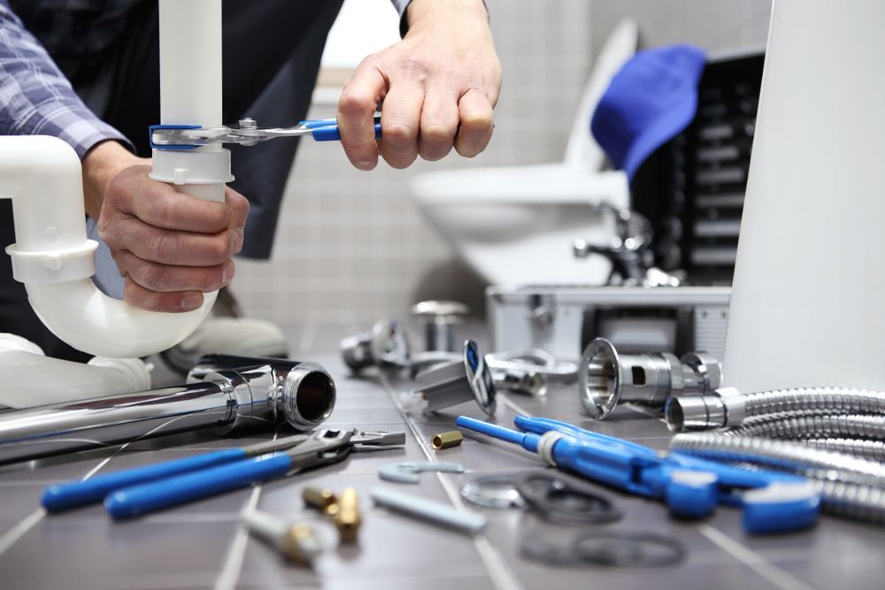 Plumber using wrench on pipe in bathroom, surrounded by tools on floor.