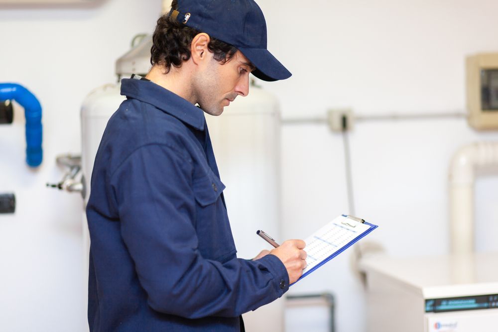 A person in a blue jumpsuit and cap writing on a clipboard in a mechanical room.