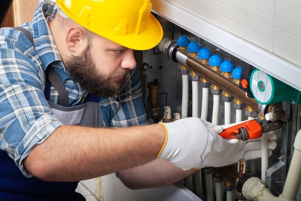 Plumber in a yellow hard hat and gloves works on pipes with a wrench.