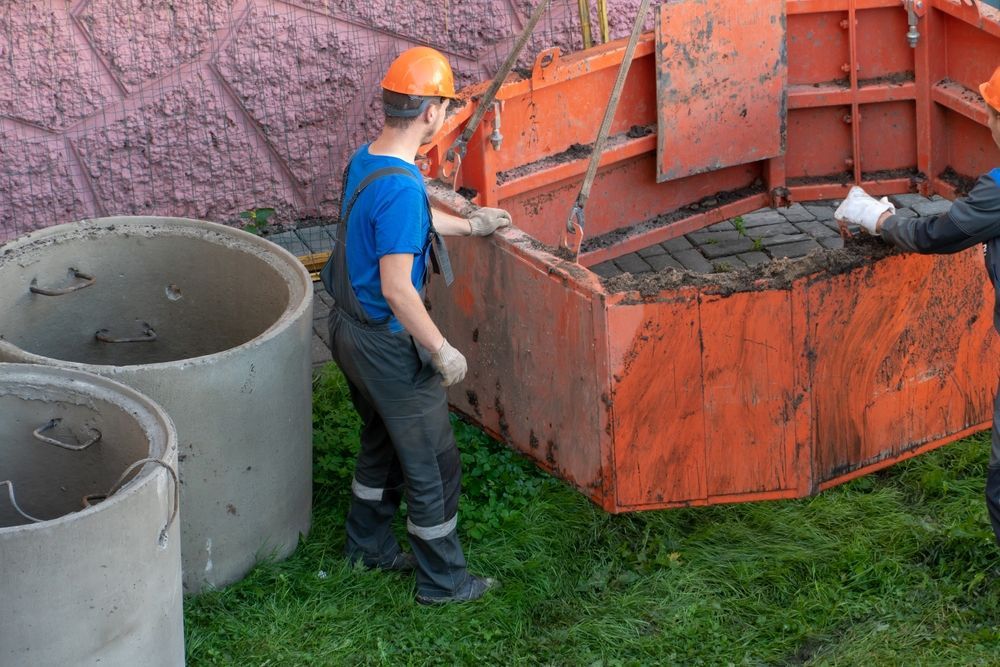 Workers in hard hats and overalls moving a large orange container near concrete rings.