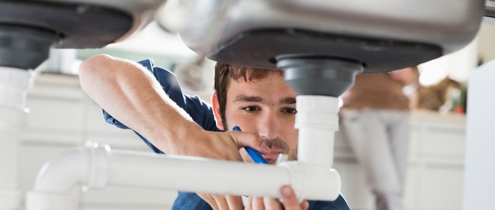 Plumber working on a sink drain in a kitchen, using a tool.