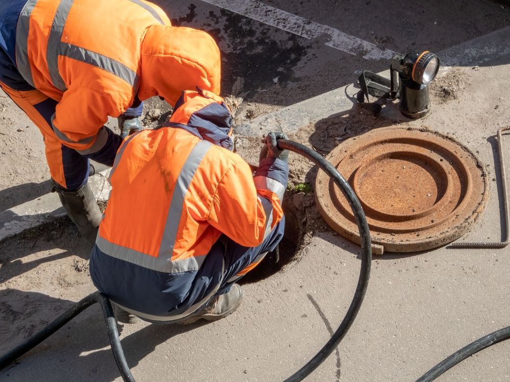 Two workers in orange vests inspect a manhole, one using a hose, outdoors.