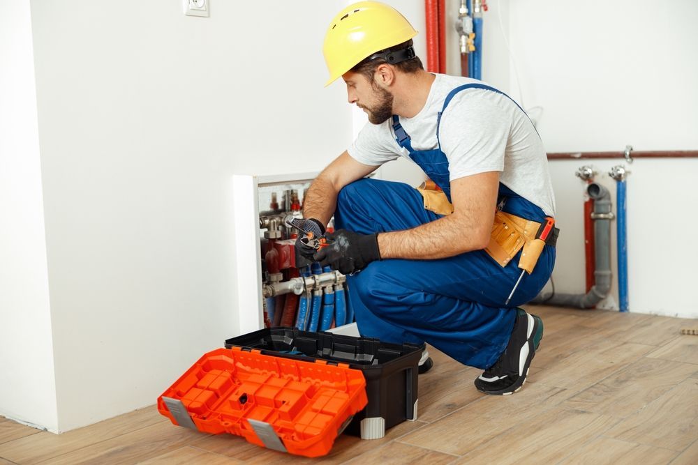 Plumber in blue overalls and yellow helmet works on pipes near an open toolbox.