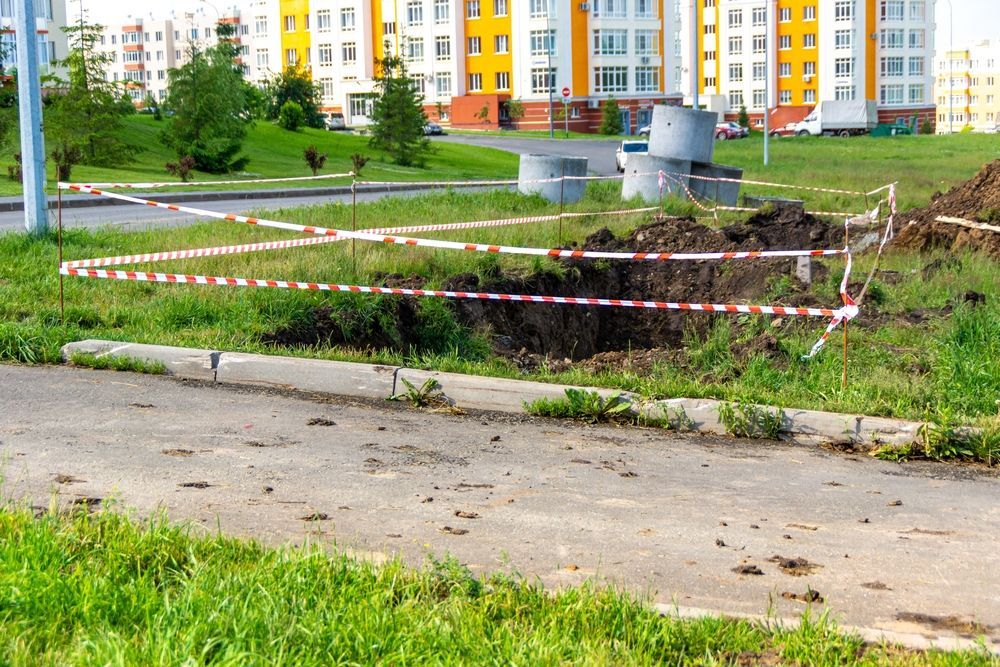 Hole dug in grass, surrounded by caution tape, near a curb and apartment buildings.