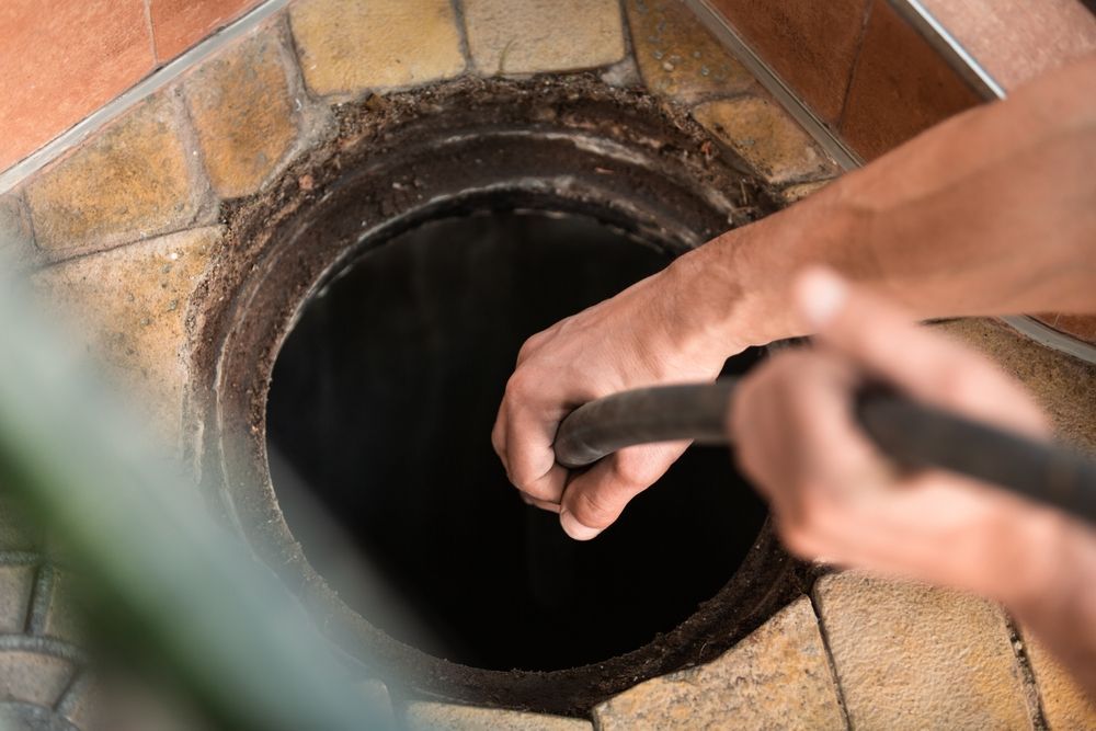 Person cleaning a sewer, holding a hose over an open manhole, surrounded by brick tiles.
