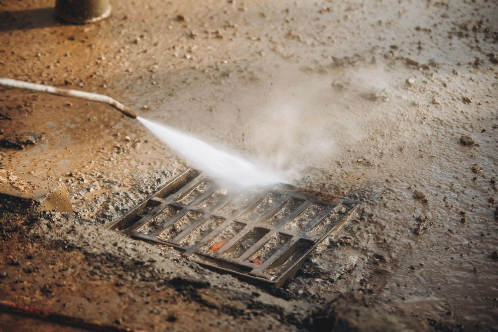 High-pressure water cleaning a dirty metal drain grate on a concrete surface.