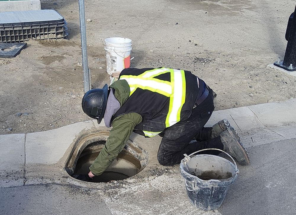 Person in safety vest kneeling over an open drain, inspecting it with a bucket nearby.
