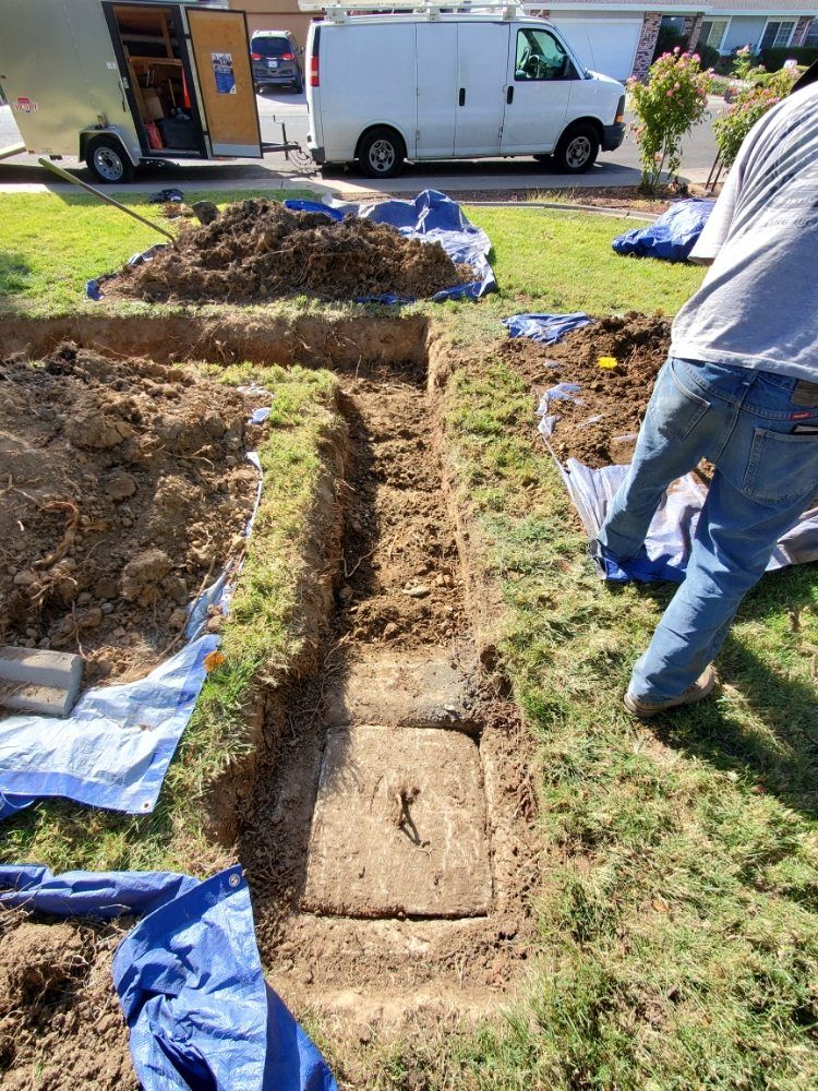 A person digging a trench around a concrete square in a grassy yard. Dirt and blue tarps surround the work.