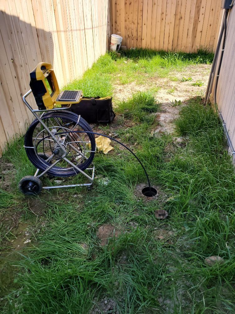A black drain camera descends into a hole in a grassy yard, near a fence, with the equipment on a cart.