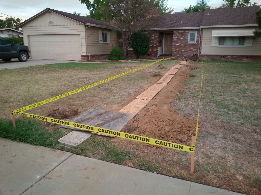 A residential yard under construction, marked with caution tape, with a trench dug in the lawn.