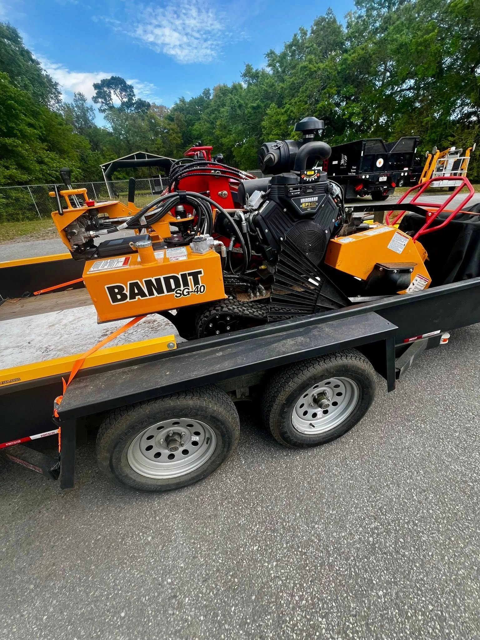 A bright orange Bandit stump grinder loaded onto a black flatbed trailer on a gravel lot under a clear blue sky.