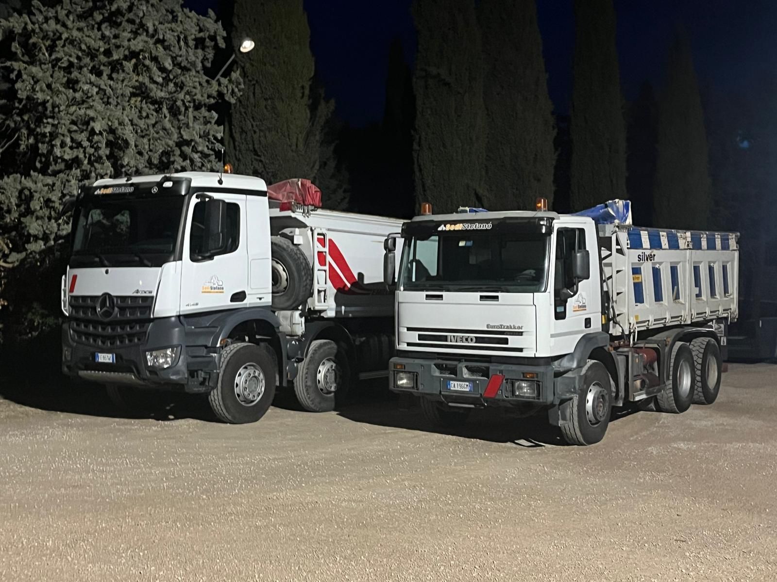 Due autocarri con cassone ribaltabile sono parcheggiati uno accanto all'altro in un piazzale ghiaioso.