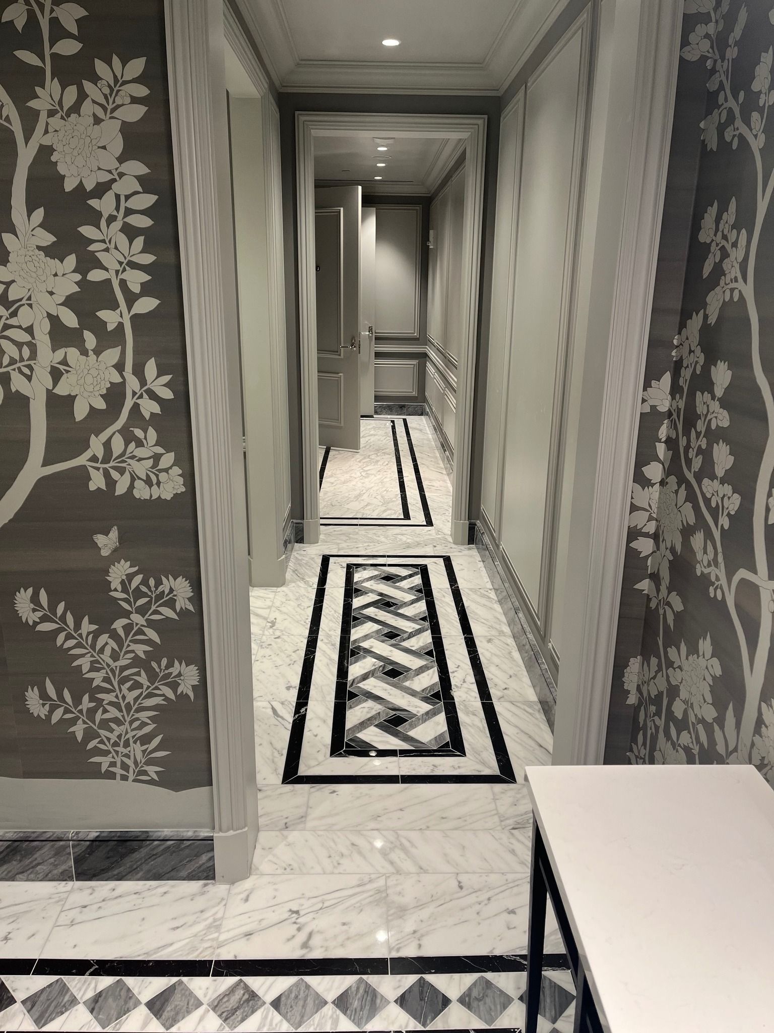 Narrow hotel hallway with patterned carpet and floral wallpaper, leading to a distant doorway.