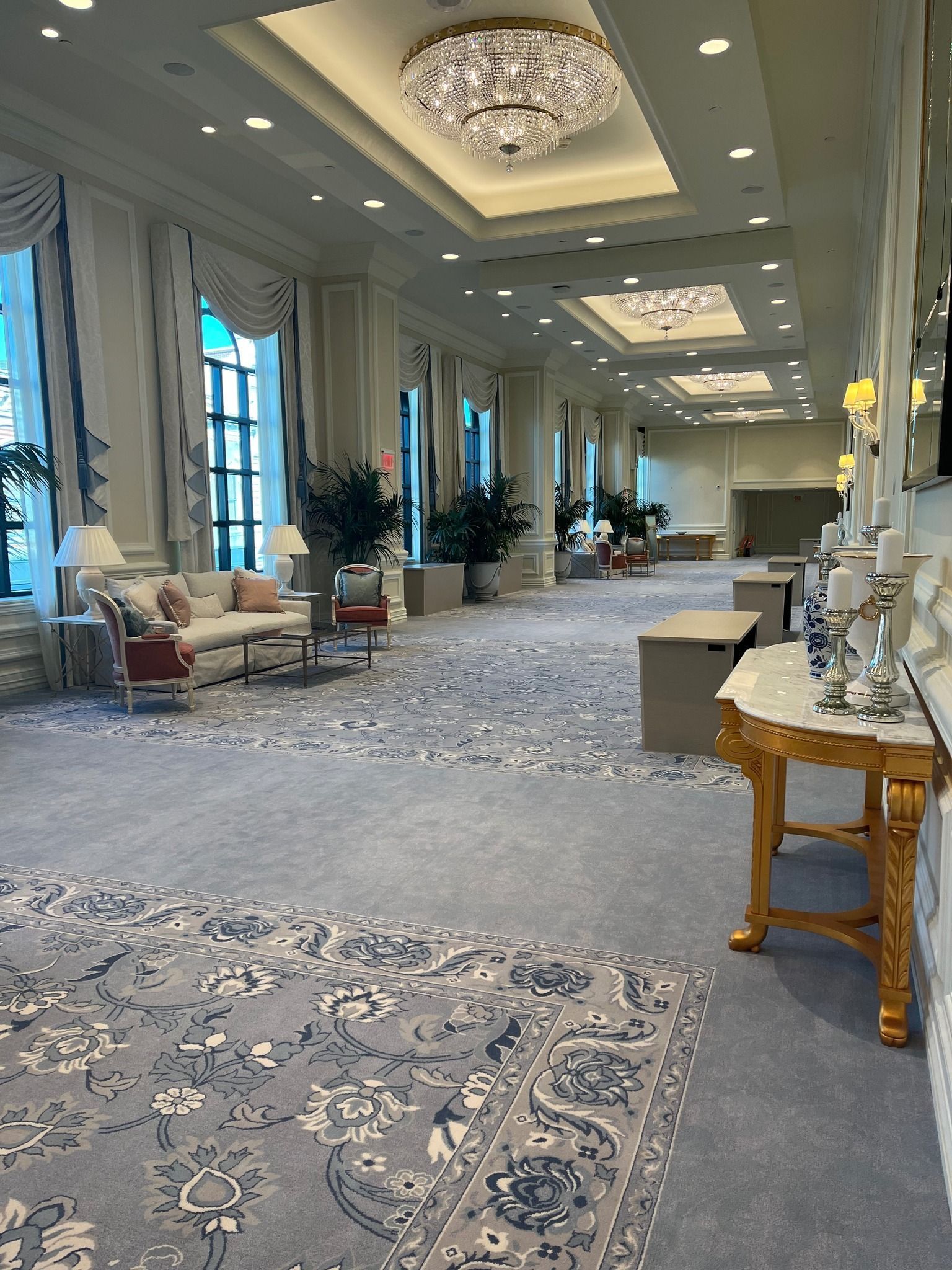 Elegant hotel ballroom with chandeliers, patterned carpet, tall windows, and seating along the walls