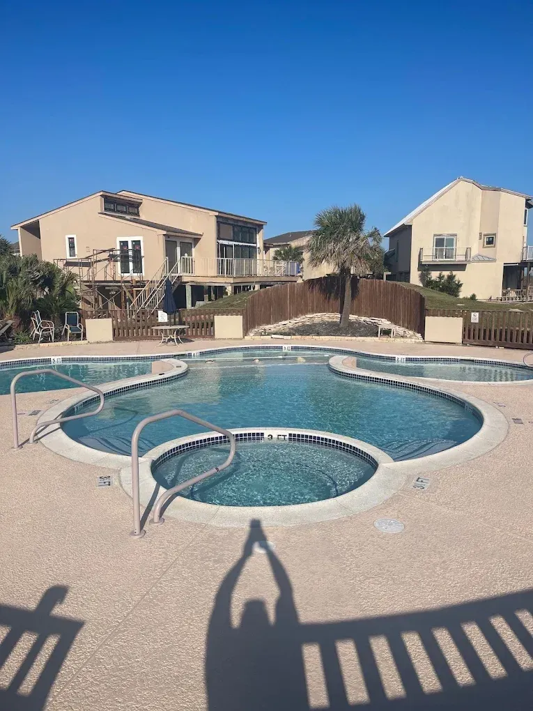 Pool with connected hot tubs in front of beige buildings on a sunny day. Shadow of person taking picture.