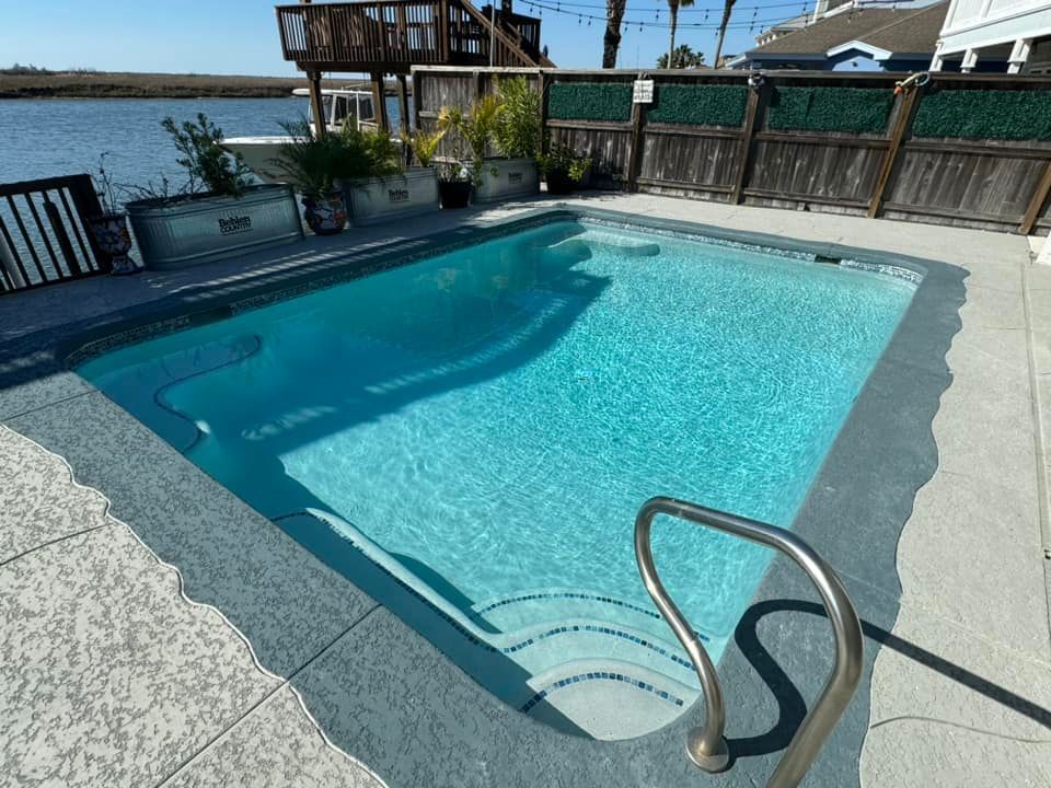 Pool with steps and handrail; blue water, concrete patio, waterfront view.