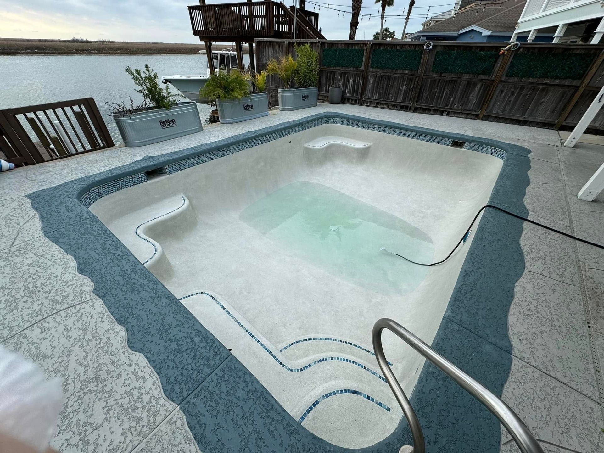 Empty rectangular swimming pool with blue tile border, steps, and handrail, outside near water.
