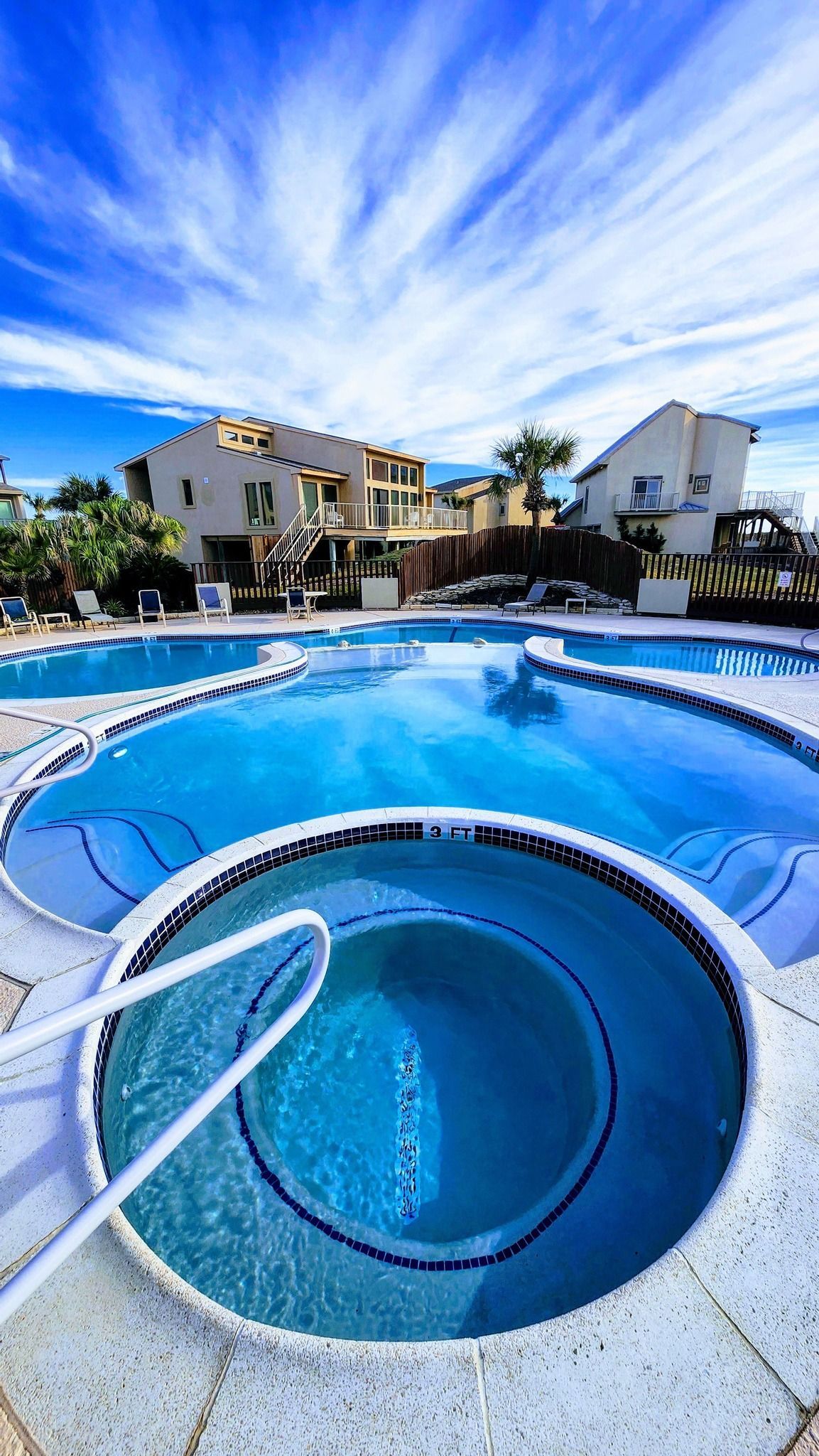 Pool area with blue water, white tiles, and buildings under a bright, cloudy sky.