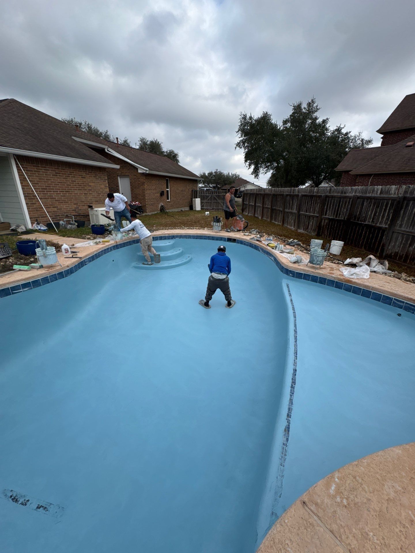 Workers painting a drained, blue-painted swimming pool on a cloudy day. Houses and a fence surround the pool.