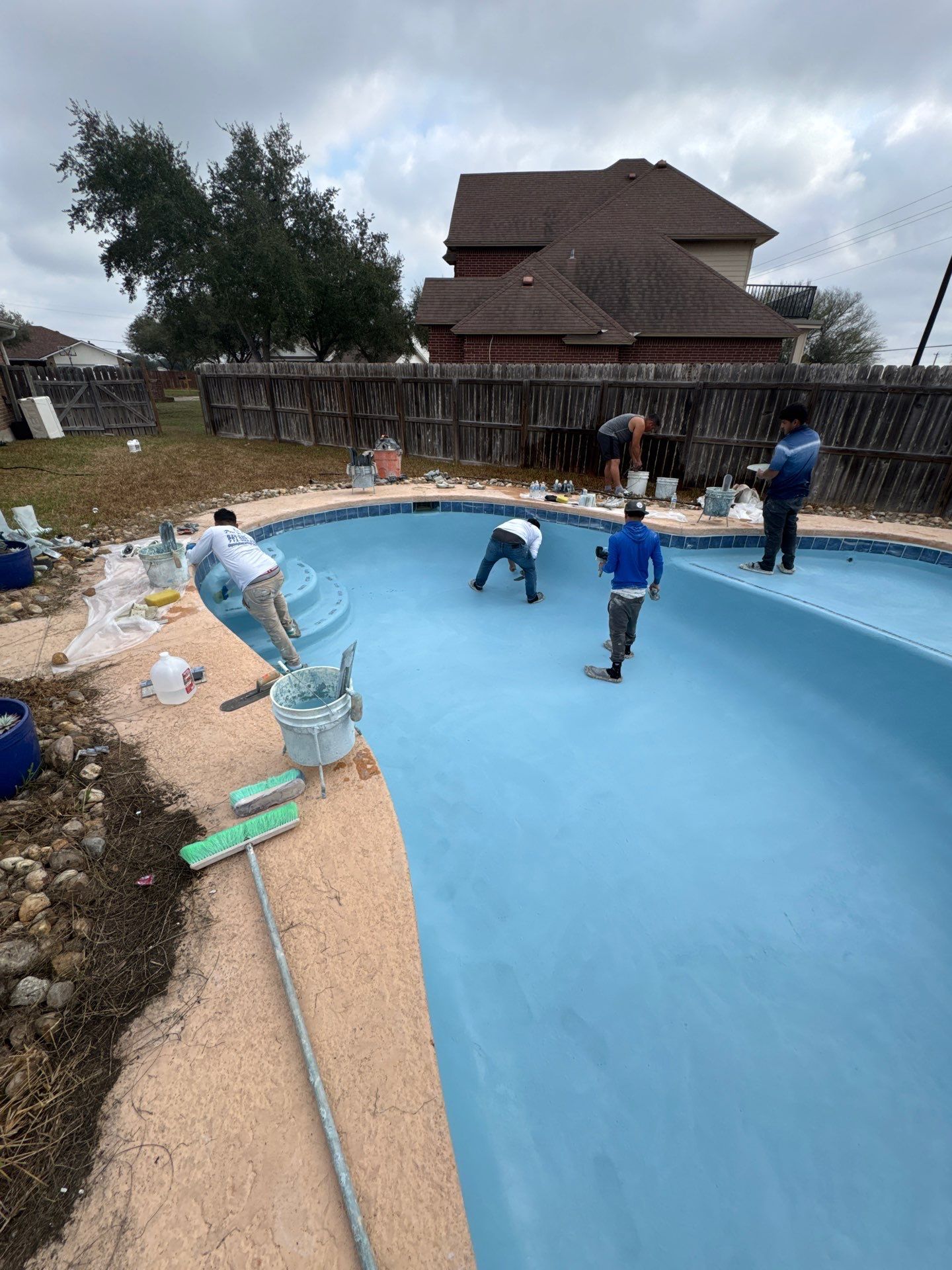Workers applying blue paint to a pool interior; outdoor setting with fence and house visible.