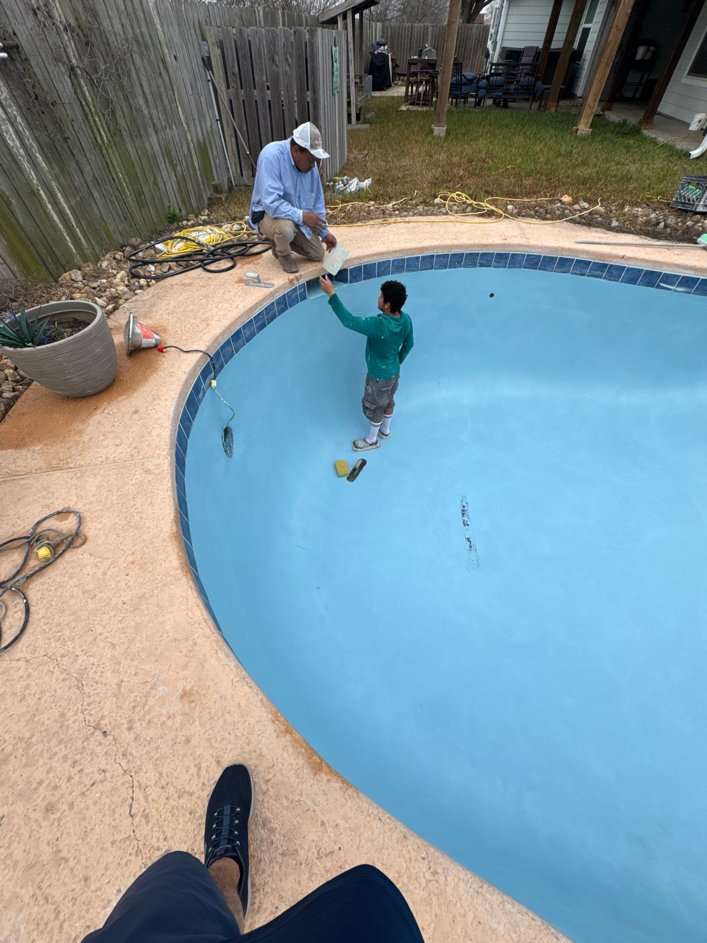 Two people inside and outside a drained pool. One paints the pool bottom; the other watches.