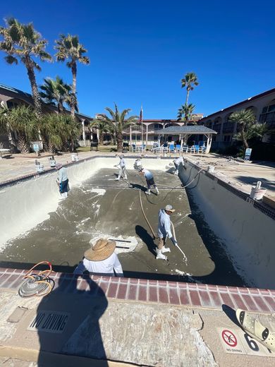 Workers resurfacing a drained outdoor pool at a resort. Palm trees and blue sky overhead.