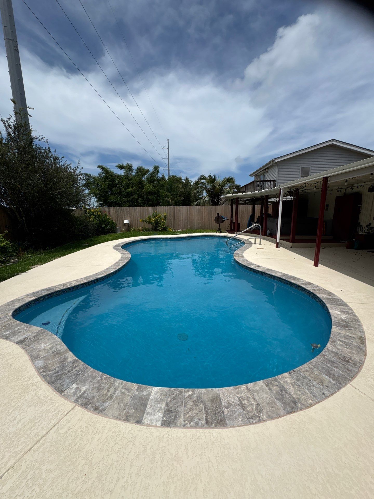 Blue, freeform pool surrounded by light concrete. House and cloudy sky in background.