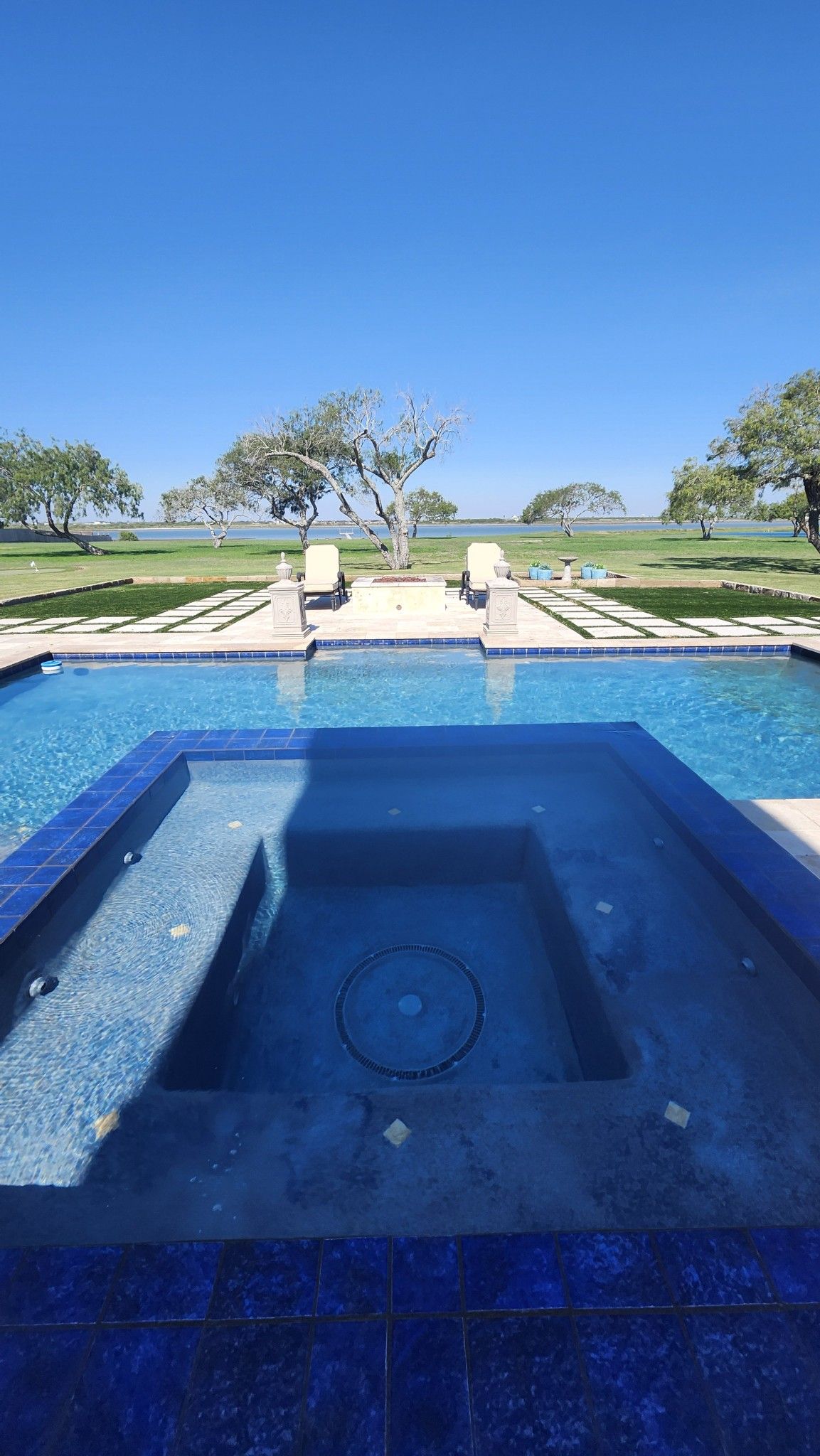 Swimming pool with jacuzzi, blue tiles, lawn, trees, and a clear blue sky.