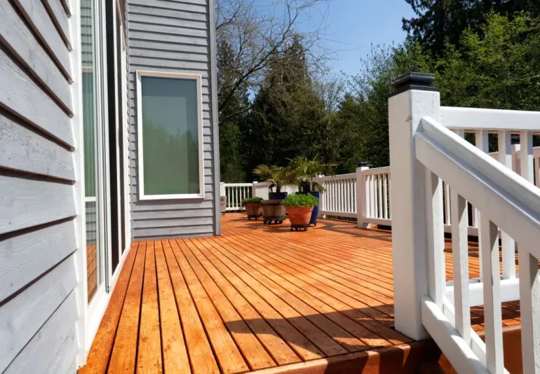 Wooden deck beside a house, with white railing, gray siding, and potted plants in sunlight.