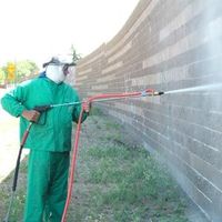 Worker in green protective gear spraying a brick wall with a pressure washer