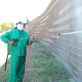 Worker in green protective gear spraying a brick wall with a pressure washer