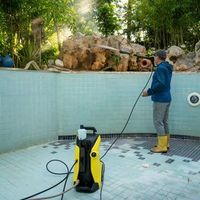 Person pressure-washing an empty pool with a yellow washer and hose, surrounded by trees.