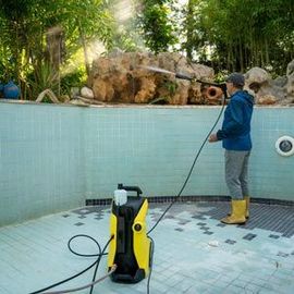 Person pressure-washing an empty pool with a yellow washer and hose, surrounded by trees.