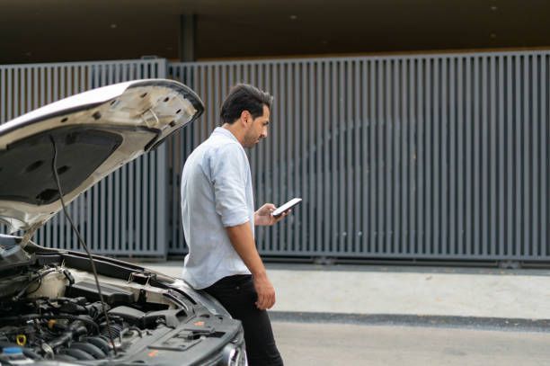 Man with car trouble on side of road looking at his phone, grey metal gate in the background.