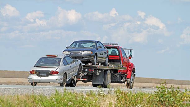 A red tow truck carrying two damaged cars on a highway against a cloudy sky.