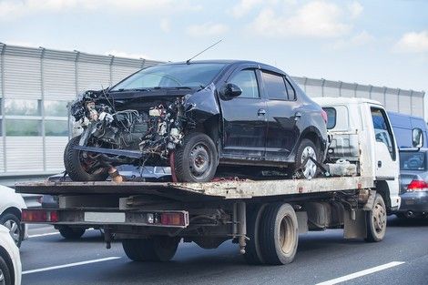 Damaged black car on a flatbed tow truck on a highway, likely after a collision.
