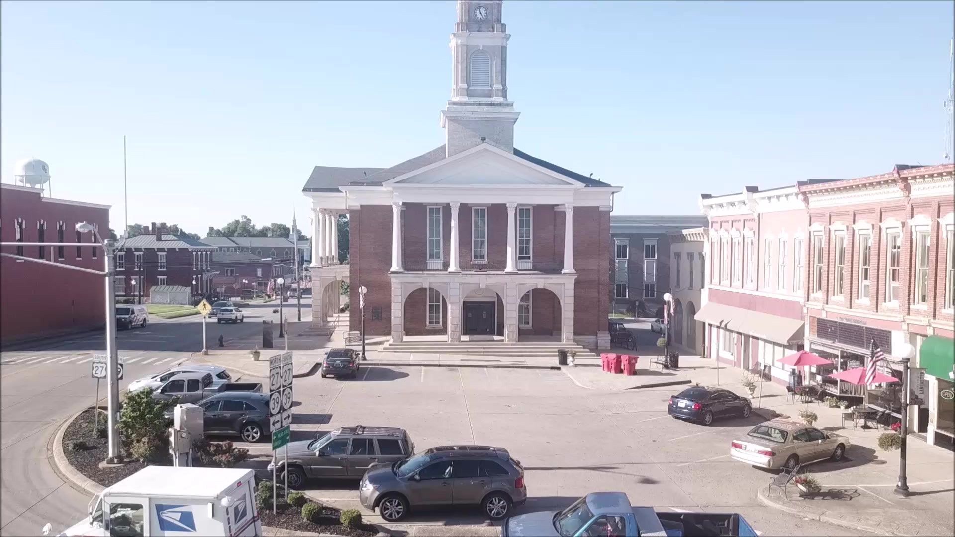 A large building with a clock tower is in the middle of a city with cars parked in front of it.