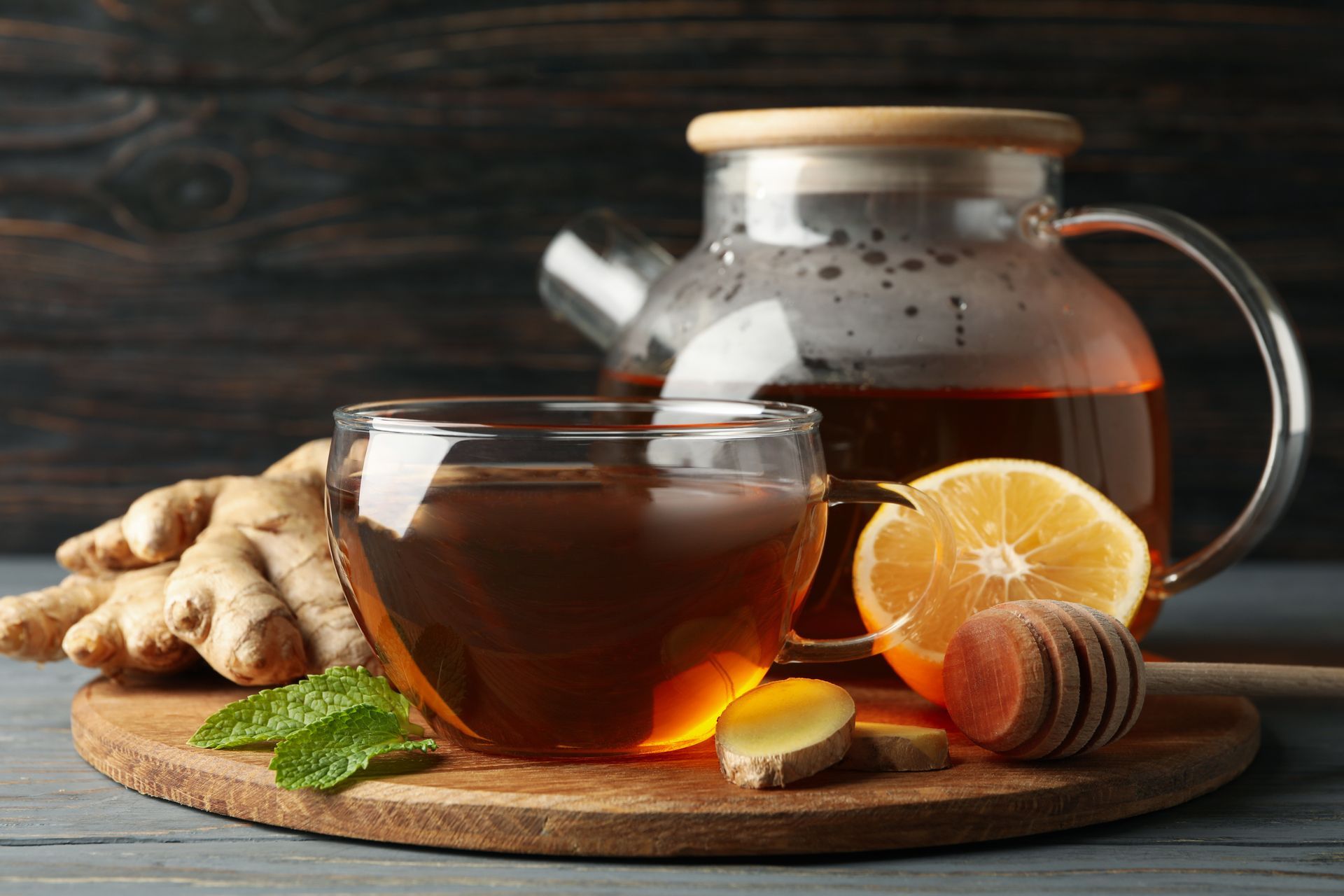 A cup of hot tea on a wooden board with ginger, lemon slices, mint, and a honey dipper.