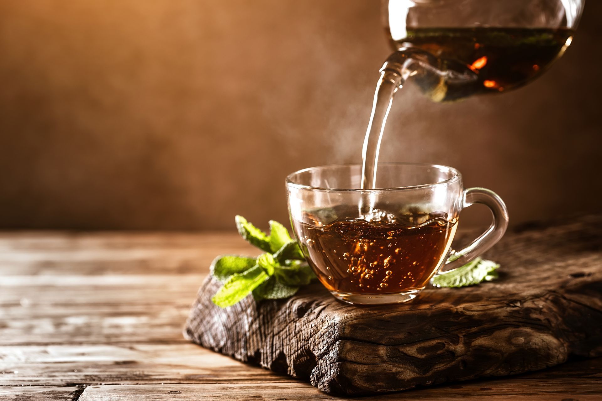 Hot tea being poured into a clear glass cup on a rustic wooden surface with fresh mint.