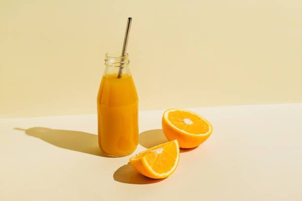 Orange juice in a glass bottle with a metal straw, sliced oranges beside it on a cream background.