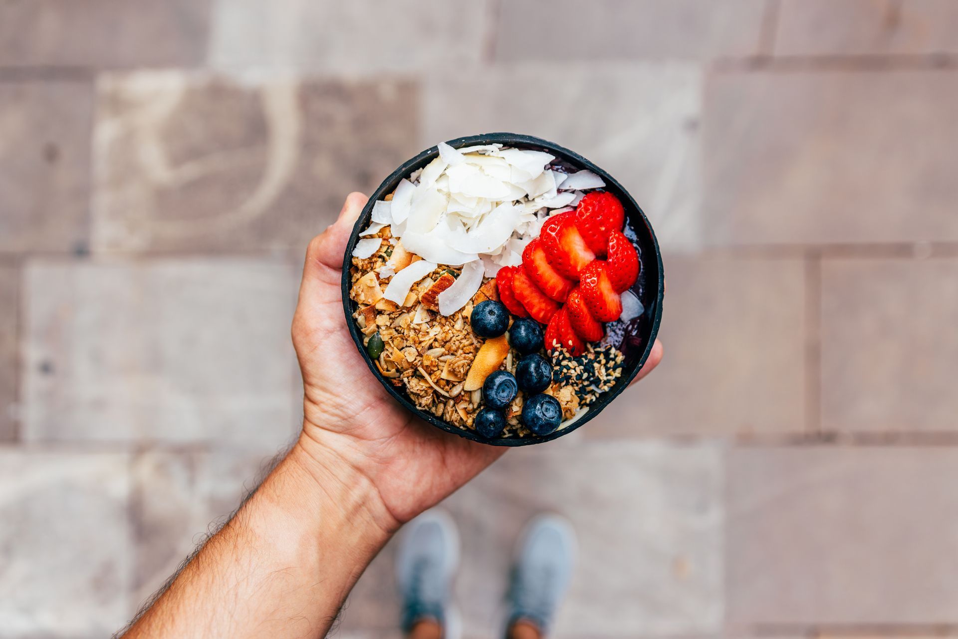 Hand holding a bowl of granola topped with coconut flakes, strawberries, and blueberries.