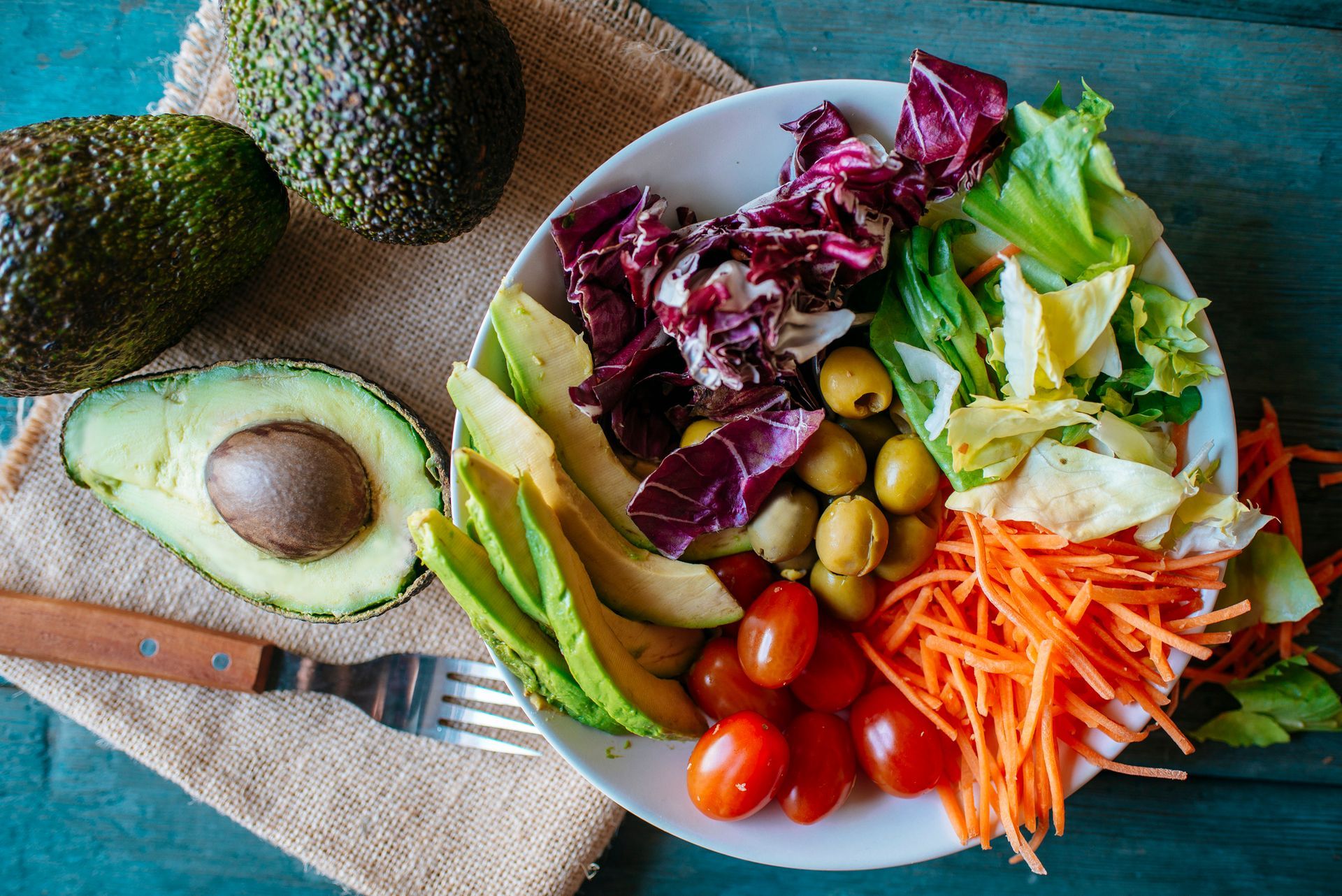 Fresh salad with avocado, tomato, carrot, olives, and lettuce on rustic blue wood.