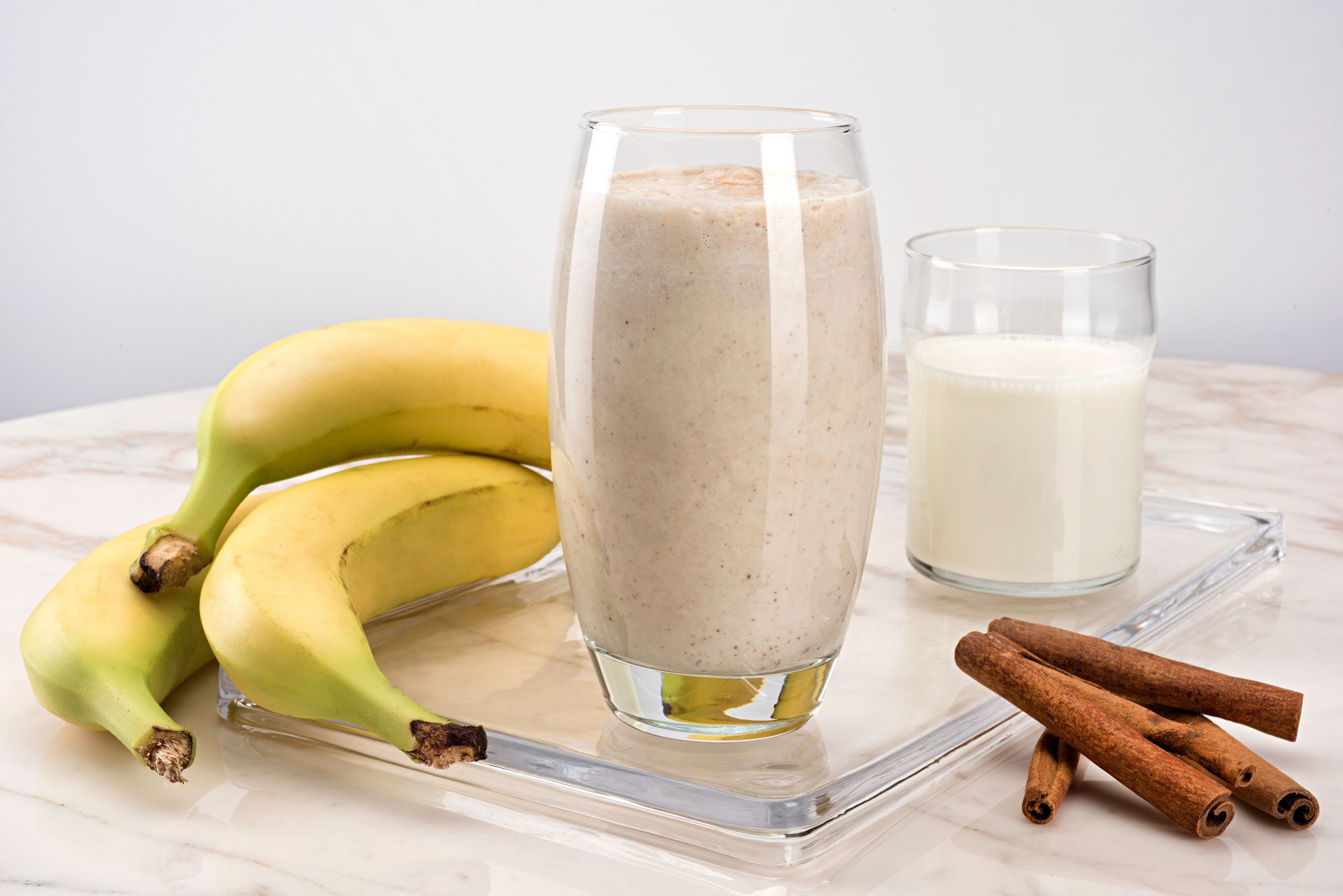 Banana smoothie in tall glass with bananas, milk, and cinnamon sticks on a tray.
