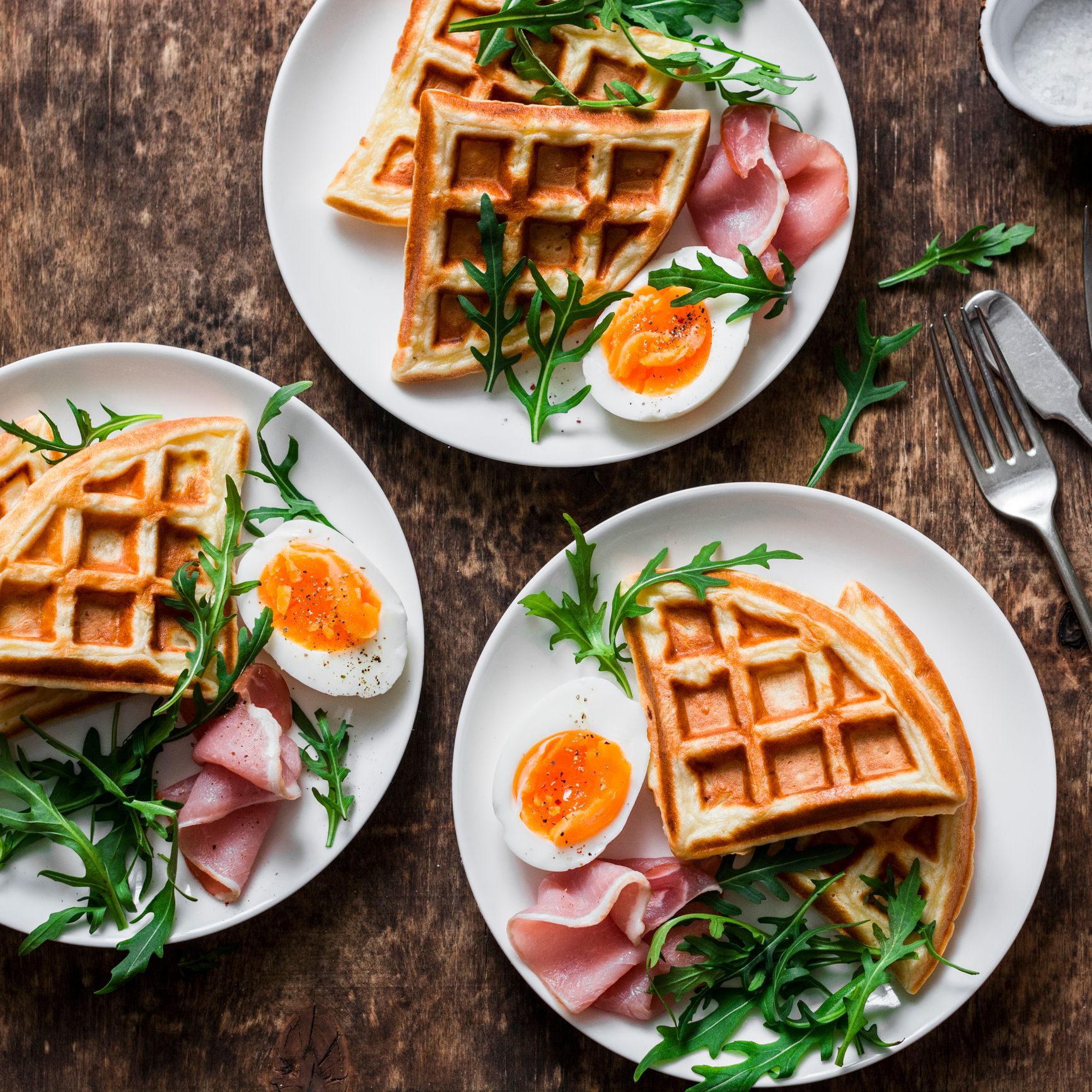 Three plates of waffles, ham, arugula, and a halved boiled egg on a wooden table.