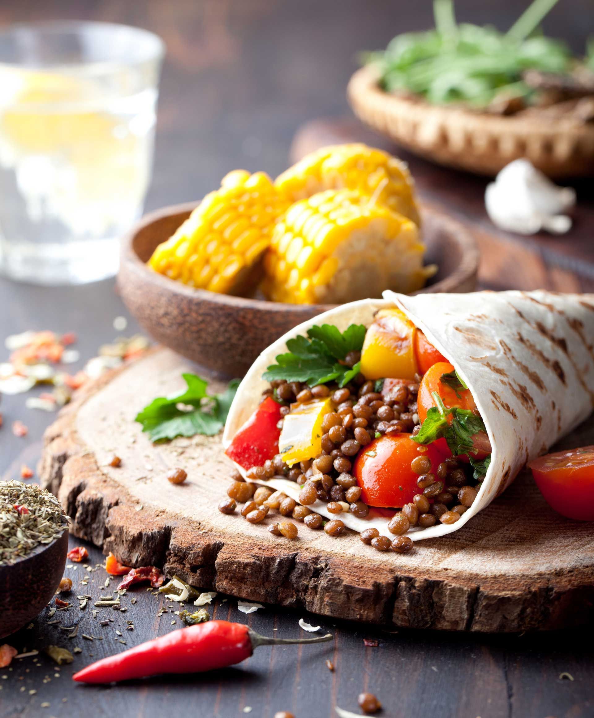 Vegan tortilla wrap filled with lentils and grilled vegetables, in front of a bowl of corn on the cob.